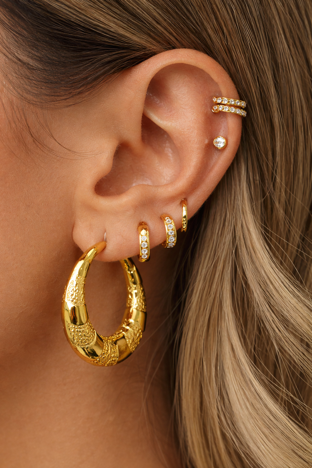 Close-up of a woman’s ear styled with multiple gold earrings, featuring a bold textured gold hoop as the main piece alongside smaller studs and hoops for a layered, modern look.